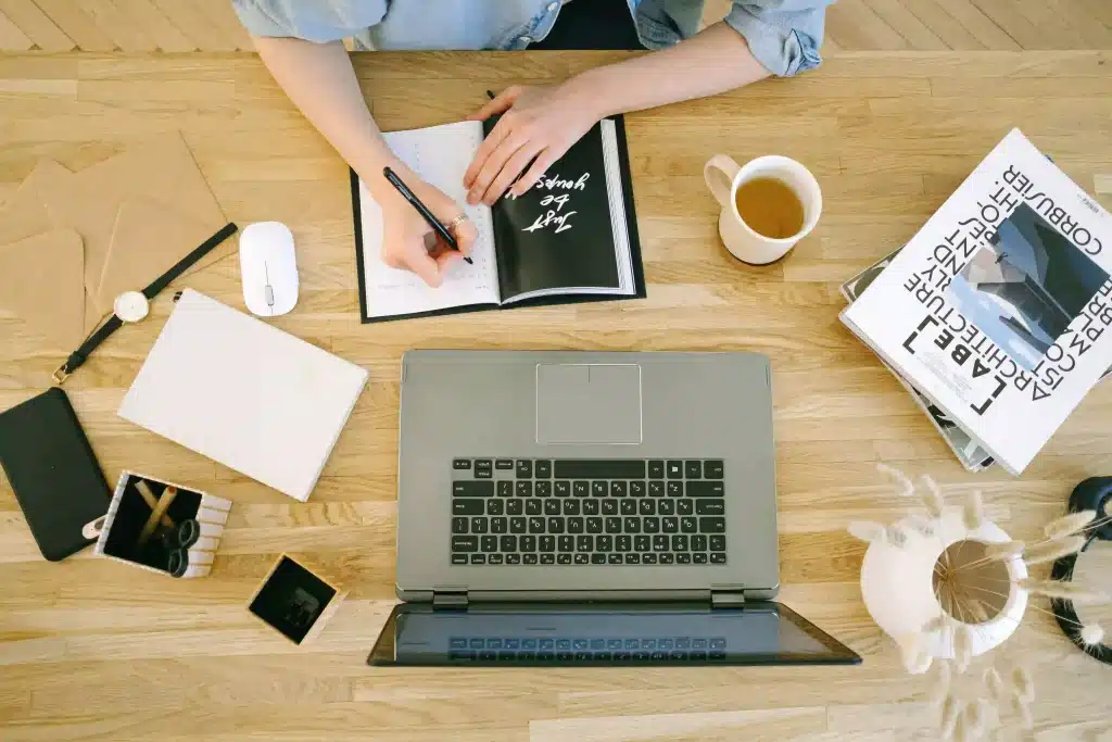 A laptop on a table and a person scribbling on a notebook